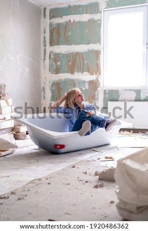 Renovation apartment. Creative story young happy woman sits in bathtub in the middle of the room. Empty walls, repairs house with their own hands. Royalty-Free Stock Photo #1920486362