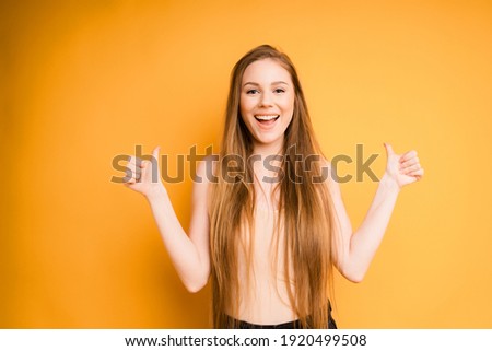 Caucasian girl with long hair smiling at the camera, showing thumb, isolated on yellow background. Positive concept
