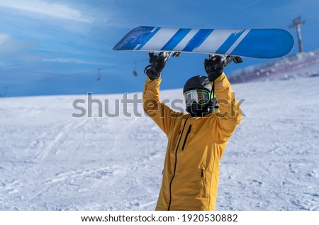 A snowboarder in a yellow jacket holds the snowboard over his head at the ski resort