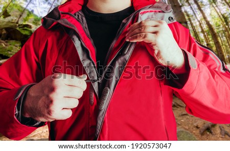 Photo of a male person putting on red travel hiking jacket in summer woods background.
