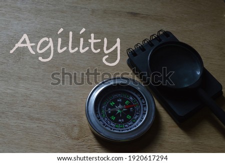 Top angle view of notebook, magnetic compass and magnifying glass over wooden background written with text AGILITY.