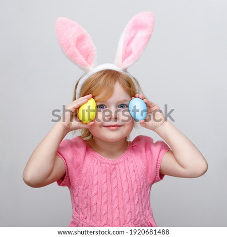 Beautiful smiling little girl wearing Easter bunny ears and holding Easter colored eggs on white wall background.