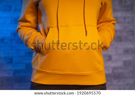 Close up of woman's hands of unrecognizable person in pockets of yellow hoodie on gray brick background. Front view on body part of young female in sweatshirt