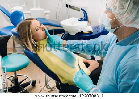 a dentist wearing a protective mask sits down next to the patient and covers his mouth with a finger. Royalty-Free Stock Photo #1920708311