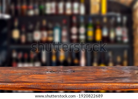 Wooden board on a background of bottles with alcohol. Old bar counter as layout for design. Workpiece for design. Empty place to advertise products. Blurred interior of the bar in the background. Royalty-Free Stock Photo #1920822689