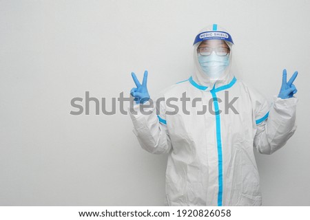 Frontliner in happy mood showing peace sign in white and blue stripe jumpsuit wearing transparent protective shield handling Covid-19 situation with white clean background and copyspace. 