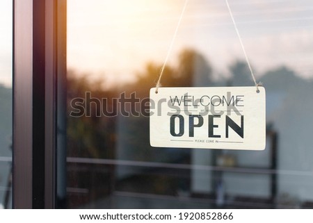 A board hung open in front of the glass door of the coffee shop. The soft light of the morning sun is bright as the background. Reflection from nature tone filter effect color style, sky, and trees.
