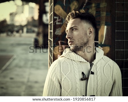Handsome young man in city setting, with industrial site around him, looking away, serious expression