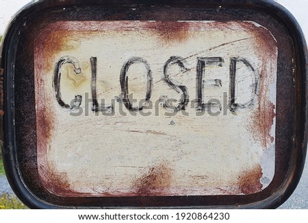A vintage metal sign with the word "Closed" written in black ink. On a pale yellow background.