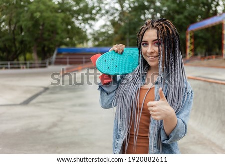 Cheerful extraordinary millennial woman with bright makeup and African braids holding penny board in a skatepark