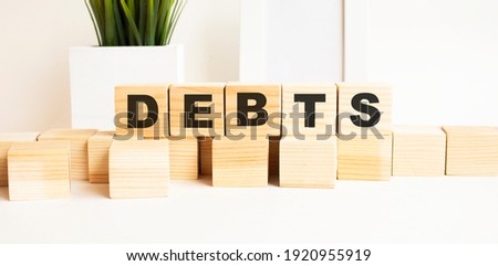 Wooden cubes with letters on a white table. The word is DEBTS. White background with photo frame, house plant.