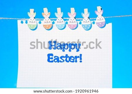 Happy Easter greeting card. On a rope hang clothespins in the shape of Easter eggs-rabbits and hold a sheet from a notebook with the inscription happy Easter.