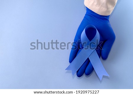 Hand in doctor's glove holding blue ribbon on blue background with copy space. Colorectal Cancer Awareness. Colon cancer in an elderly person. World Diabetes Day.