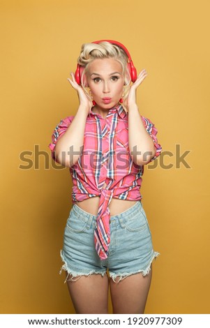 Pretty pinup woman listening music with headphones on bright yellow background. Pretty retro model with red lips makeup and vintage fashion hairstyle