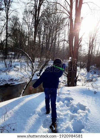 picture of winter on the banks of a small river, a boy in a jacket and a hat throws a snowball with gusto