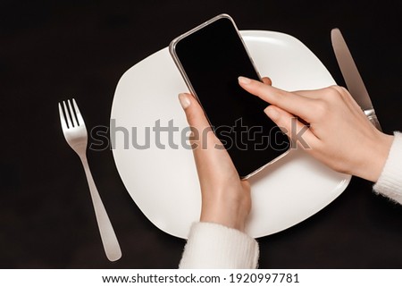 Smart phone screen on white plate, fork and knife, black background table. Take away restaurant meal online, food delivery concept.