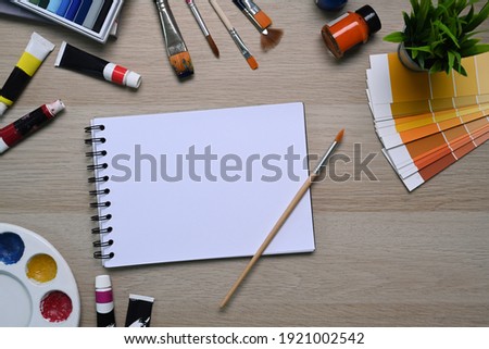 Artist workspace with empty notebook surrounded by color swatches, paintbrushes and other various equipment on wooden background.