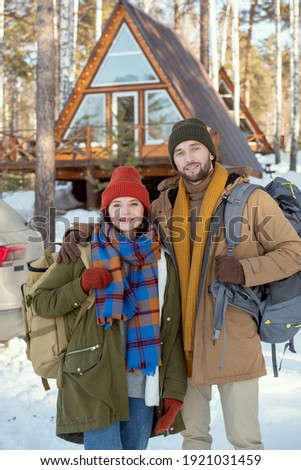 Young affectionate couple with big backpacks standing in front of camera against their car and country house and looking at you outside