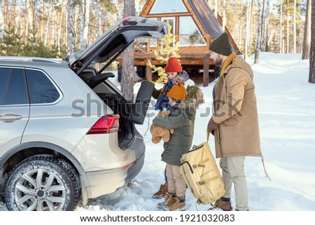 Contemporary family of three in warm winterwear standing by back of the car and putting their buggage into trunk while going to travel