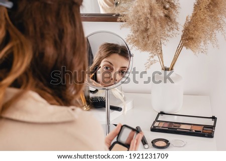 Young woman applying make-up to her face, using black eyelliner for eyes and looking at the round mirror. Feminine beauty routine at home. Selective focus
