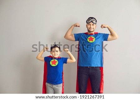 Powerful father with son in superhero costume showing muscles on gray background. Little boy wants to have the same biceps as his father. Family is dressed in red cloaks and pilot glasses. Banner. Royalty-Free Stock Photo #1921267391