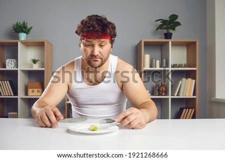 When slimming stops being healthy. Unhappy guy keeping strict diet and eating less food. Sad young man sitting at table with almost nothing on plate looking at tiny little portion of fruit for lunch Royalty-Free Stock Photo #1921268666