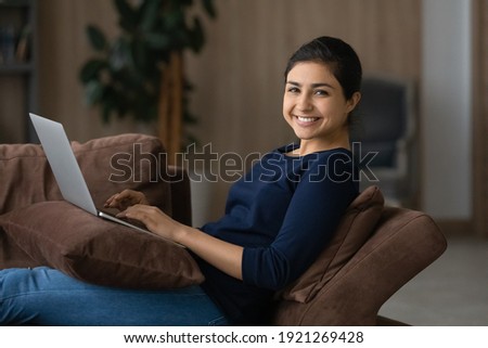 Portrait of smiling young Indian woman sit on sofa at home work online on laptop device. Happy millennial mixed race female relax on couch in living room study distant on computer. Technology concept. Royalty-Free Stock Photo #1921269428