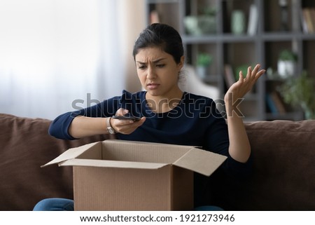 Unhappy young Indian woman frustrated by wrong order shopping online using smartphone. Upset ethnic female buyer unbox parcel buying on web confused with bad delivery service. Shipping concept. Royalty-Free Stock Photo #1921273946