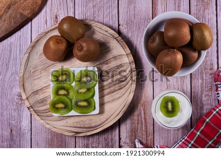 Yoghurt glass with kiwis on wooden plate. Wooden floor, red and white kitchen towel, chequered, zenithal view, kiwi cut on rectangular plate.