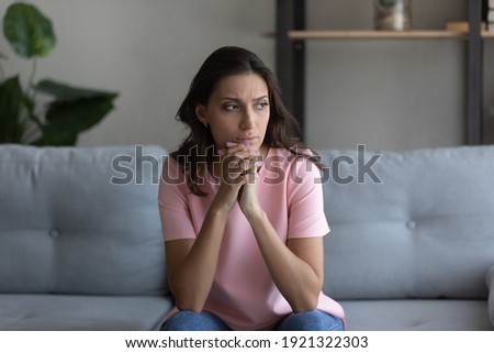 Doubtful millennial arab female sitting on couch at home lost in serious thoughts. Thoughtful young woman of indian ethnicity looking aside pondering on making choice decision solving problem in mind Royalty-Free Stock Photo #1921322303