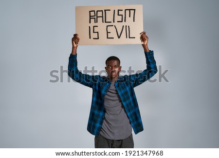 Confident young african american guy in casual clothes looking at camera, holding demonstration banner with Racism is evil text above his head, posing isolated over gray background. Front view