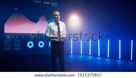 Middle aged man gesticulating and talking against LED display with financial charts during business conference