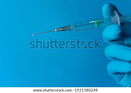 A hand with a nitrile glove appears on the right side of the image, holding an injection. On the tip of the needle, a drop of liquid is about to fall, all on a blue background with copy space. Royalty-Free Stock Photo #1921380248