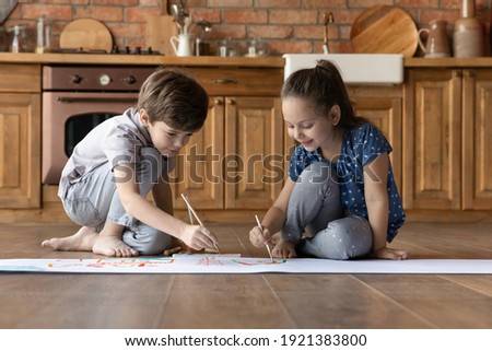 Two little kids painting colorful watercolor, sitting on warm wooden floor in kitchen, cute sister and brother playing, enjoying leisure time, engaged in creative activity at home together