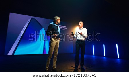 Mature businessman showing new cellphone and speaking with bearded engineer while standing against LED screen during presentation on stage Royalty-Free Stock Photo #1921387541