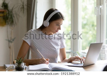 Focused young Caucasian female in headphones look at computer screen take online education course or training. Millennial woman in earphones study or work distant on laptop at home workplace. Royalty-Free Stock Photo #1921388912