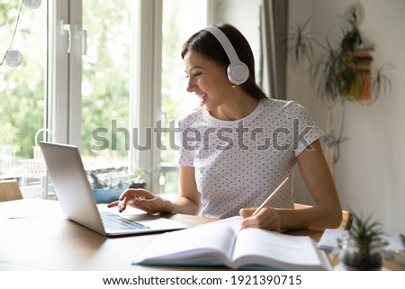 Happy millennial Caucasian female student in headphones look at laptop screen study distant from home. Smiling young woman in earphones make notes watch webinar on computer, take web course. Royalty-Free Stock Photo #1921390715