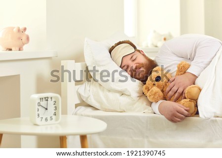 Happy grown-up man sleeping in good comfortable bed with cute teddy bear. Funny guy lying on white pillows and dreaming sweet dreams in deep sleep at 10 am in Covid-19 lockdown or on weekend morning Royalty-Free Stock Photo #1921390745