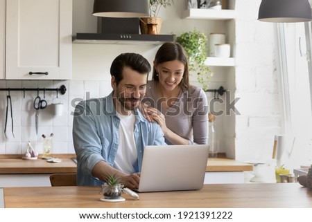 Smiling millennial couple sit at table in home kitchen using modern laptop shopping online together. Happy young Caucasian man and woman browse wireless internet on computer. Technology concept.