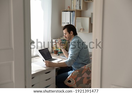 Candid view of young man sit at table at home office work on computer record audio message. Millennial male talk on loudspeaker on cellphone, activate voice assistant on cell.
