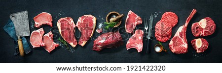 Assortment of raw cuts of meat, dry aged beef steaks and hamburger patties for grilling with seasoning and utensils on dark rustic board Royalty-Free Stock Photo #1921422320