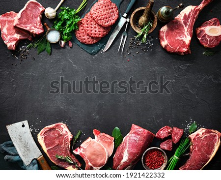 Assortment of raw cuts of meat, dry aged beef steaks and hamburger patties for grilling with seasoning and utensils on dark rustic board Royalty-Free Stock Photo #1921422332