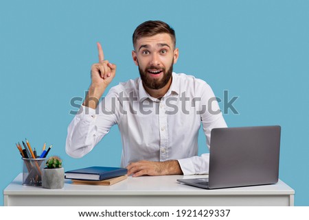 Millennial Caucasian office worker having creative idea, gesturing eureka at workplace on blue studio background. Young man finding solution to problem, sitting at his desk with laptop