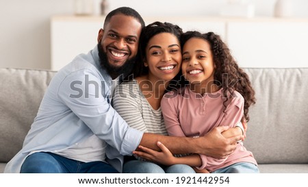 Portrait of cheerful African American family of three hugging sitting on the couch at home, posing for photo and looking at camera. Smiling young bearded man embracing his loving wife and daughter