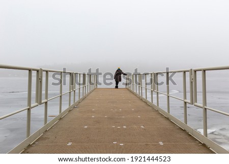 White metal pier with a wooden floor standing on the edge of the dam. A man stands at the end of the pier and looks into a karajina full of fog.