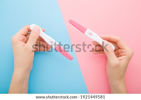 Young adult woman hands holding pregnancy tests with one stripe and two stripes on light blue pink table background. Pastel color. Negative and positive result. Closeup. Point of view shot. 