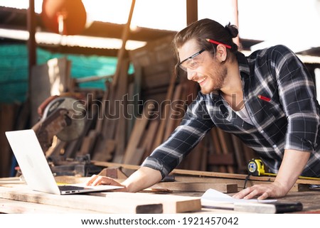 Young woodworker with beard leaning over workbench in his large workshop full of carpentry equipment working online with laptop