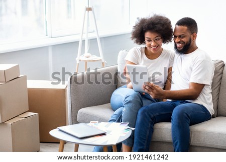 Real Estate Application. Cheerful African Spouses Using Tablet Computer Searching Apartment For Rent Online Sitting Together On Sofa At Home. House Hunting And Relocation Concept