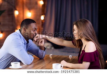 Happy Interracial Couple Flirting During Romantic Date In Restaurant, Loving African American Guy Kissing Hand Of His White Girlfriend, Multicultural Lovers Looking At Each Other, Side View