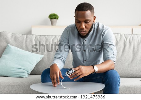 African Guy With Pulse Oximeter Clip On Hand Measuring Level Of Oxygen Saturation Sitting On Couch At Home. Ox Measurement Device, Pulseoxymetry Machine Concept. Selective Focus Royalty-Free Stock Photo #1921475936